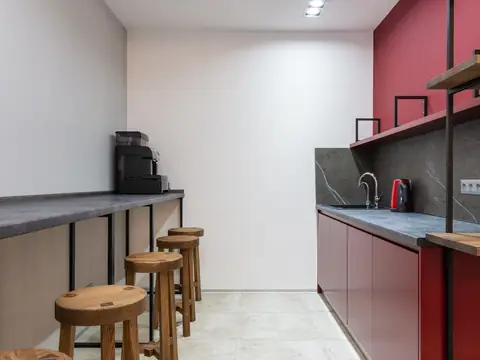 A small kitchen in a workspace with red cabinets, a sink, a kettle, a coffee machine on a counter and a row of wooden stools