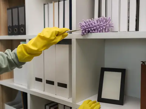 A hand in a yellow rubber glove running a purple duster along a white shelf of files and box folders