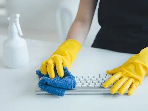 Hands in yellow rubber gloves wiping a white computer keyboard with a blue cloth on a white desk, a plain white spray bottle beside it