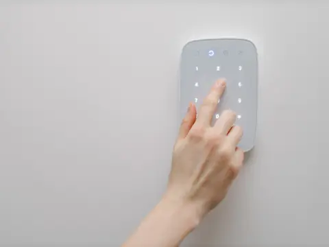 A finger pressing a number on a white alarm keypad mounted on a plain wall