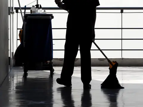 The silhouette of a person mopping a polished floor beside a janitor's cart, seen from the shoulders down against a bright railing