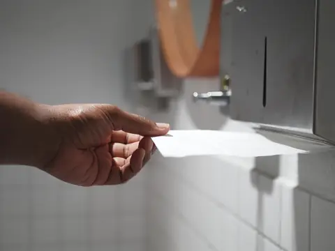 A hand pulling a paper towel from a steel wall dispenser above white tiles