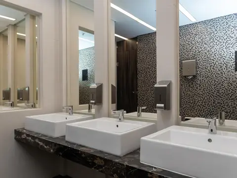 A restroom with three white basins on a dark marble counter, sensor taps, steel soap dispensers and mirrors