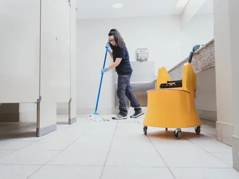 A woman in a black T-shirt and blue gloves mopping the tiled floor of a restroom beside partition stalls, a yellow mop bucket on wheels in front