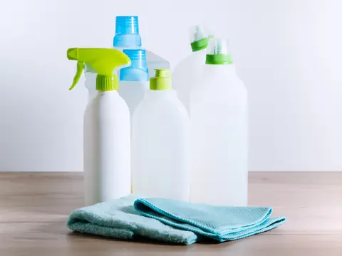 Six plain white cleaning bottles with green and blue caps, one with a spray trigger, standing behind two folded blue cloths on a wooden surface