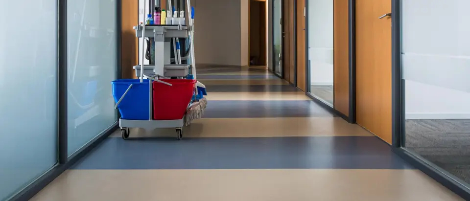 Janitor's cart with blue and red mop buckets, mops and spray bottles standing in a corridor lined with frosted glass and wood doors
