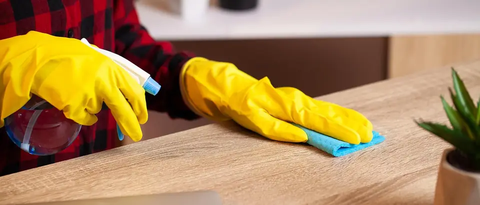 Hands in yellow rubber gloves wiping a wooden desk with a blue cloth, holding a spray bottle in the other hand